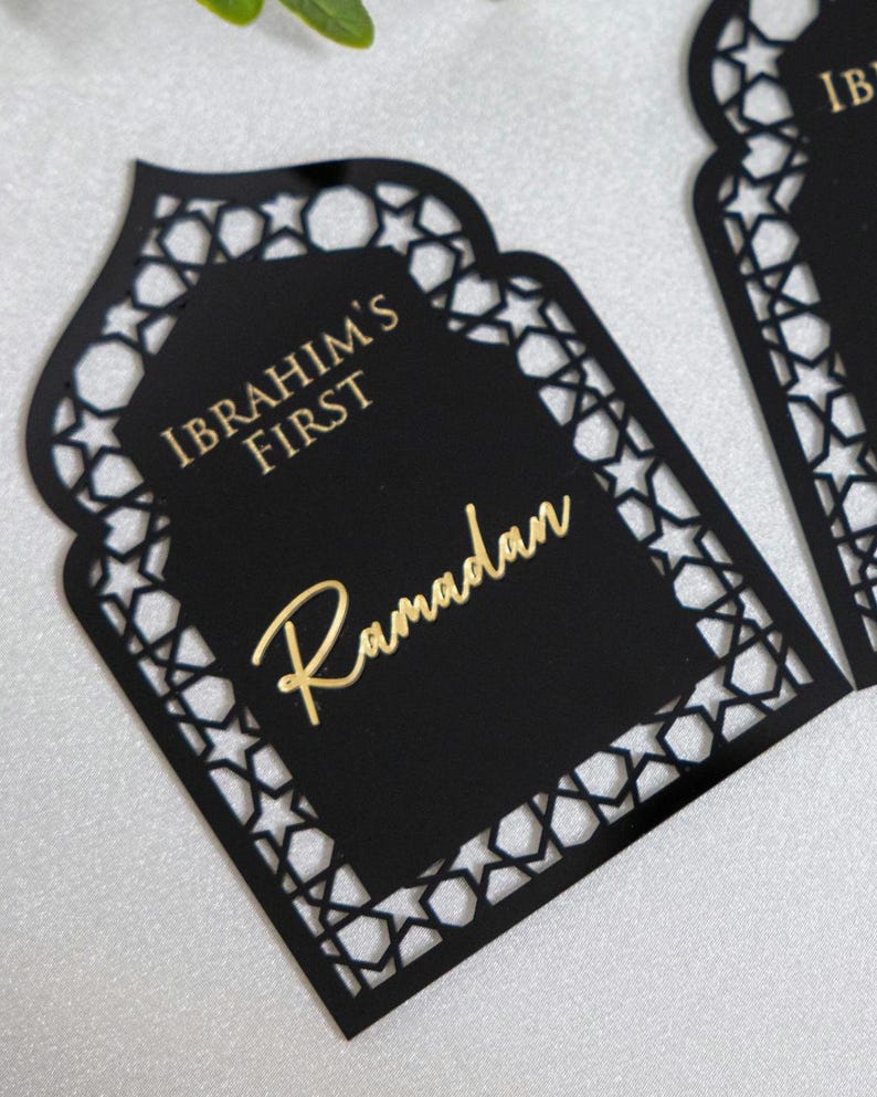 May include: Black laser-cut Ramadan decoration with a decorative star pattern border. The center features the text "IBRAHIM'S FIRST" and "Ramadan" in gold lettering. The decoration is in a unique arch shape.