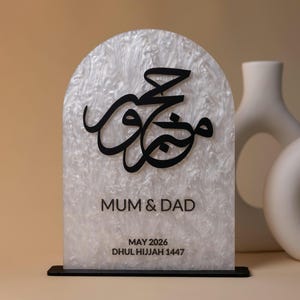 May include: A decorative sign with a white, marbled background and a black base. The sign features black Arabic calligraphy and the words "MUM & DAD" along with the date "MAY 2026" and "DHUL HIJJAH 1447".