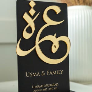 May include: A black acrylic plaque with gold Arabic calligraphy and the words "USMA & FAMILY". The plaque also includes the text "UMRAH MUBARAK AUGUST 2025 | 1447 AH". The plaque is displayed on a black base.