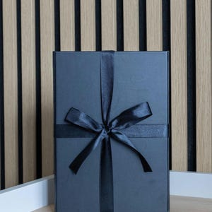 May include: A dark gray gift box tied with a black satin ribbon and bow. The box is rectangular and sits on a light wood surface, with a striped wood panel background.
