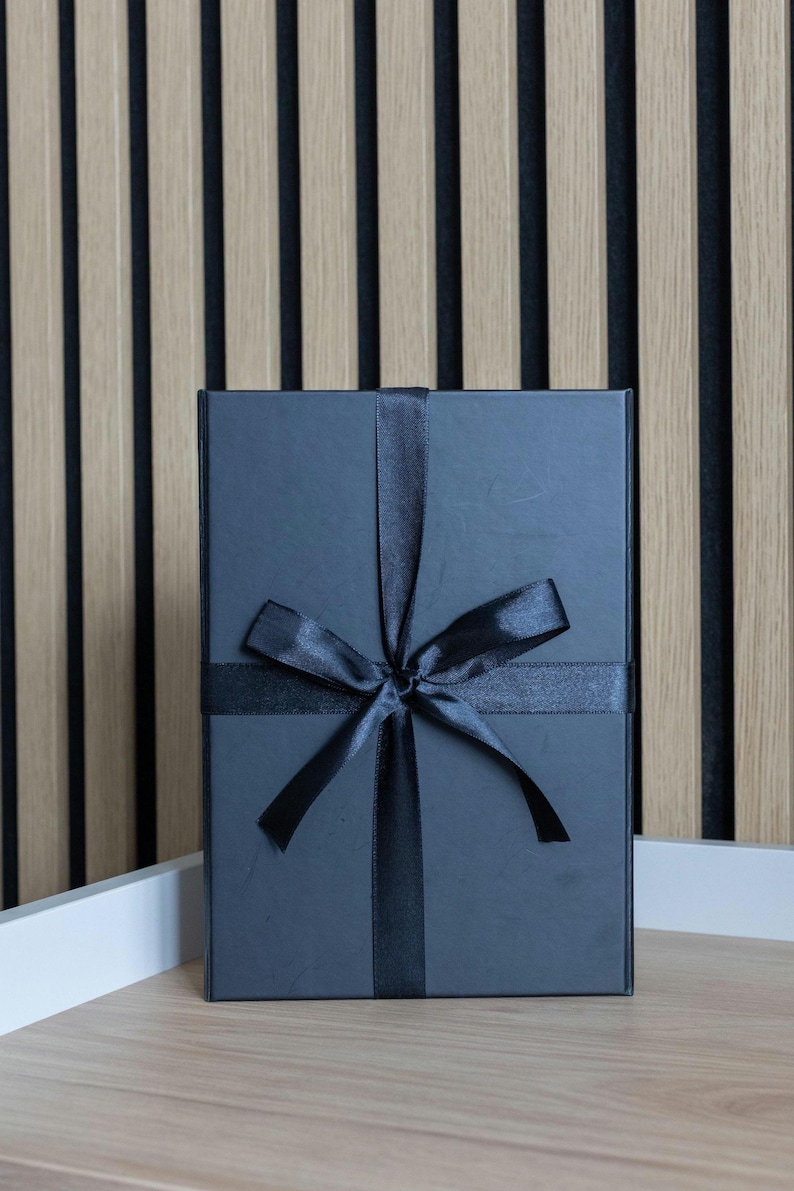 May include: A dark gray gift box tied with a black satin ribbon and bow. The box is rectangular and sits on a light wood surface, with a striped wood panel background.