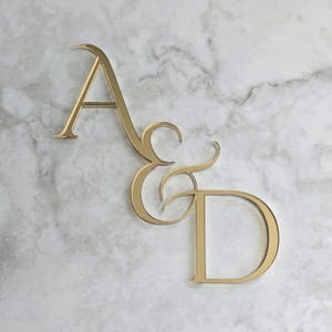 May include: A gold-coloured metal monogram displaying the letters "A", "&", and "D". The letters are in a cursive style, set against a white and grey marble background. This monogram is suitable for wedding decorations or personalised presents.
