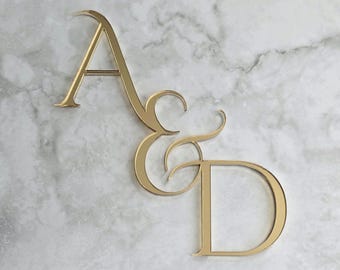 May include: Gold-colored metal monogram featuring the letters "A", "&", and "D". The letters are in a cursive style and are displayed against a white and gray marble background. The monogram is likely used for wedding decor or personalized gifts.