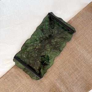 May include: A decorative green resin tray with black handles. The tray has a unique, organic shape with a wavy edge. Gold flecks are embedded in the resin, adding a touch of sparkle. The tray is designed for serving or display.