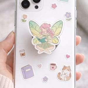 May include: Clear phone case with a fairy-themed pop socket and decorative stickers. The pop socket features a fairy reading a book. Stickers include books, flowers, hearts, coffee cups, and a cat.