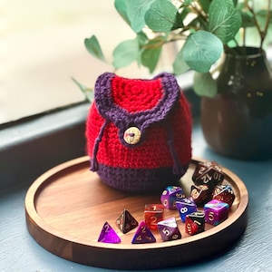 May include: A handmade red and purple crochet dice bag with a gold button closure. The bag sits on a wooden tray with a collection of colourful polyhedral dice. A decorative vase with greenery is in the background.