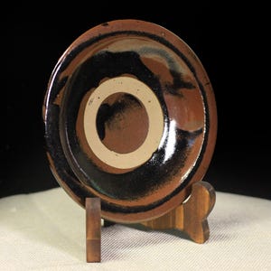 May include: A small, round ceramic bowl with a brown, black, and white color scheme. The bowl is displayed on a wooden stand. The interior features a central white circle surrounded by brown and black concentric rings.