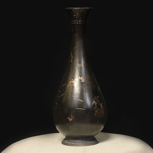 May include: A dark bronze vase with a narrow neck and a rounded body. The vase has gold-coloured decorative details, including a geometric pattern near the rim and figures. The vase is displayed on a white surface against a black backdrop.