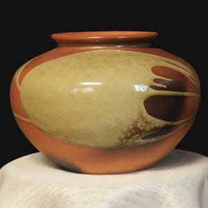 May include: A ceramic vase with a rounded form, showcasing a warm orange base and rim. The vase is decorated with a large, light tan oval design and dark brown accents, providing a striking contrast. The vase is displayed on a white cloth against a black backdrop.