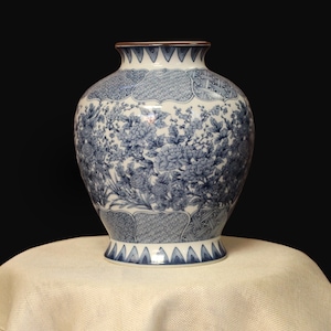 Puede incluir: Un jarrón de cerámica grande, azul y blanco, con un cuerpo ancho y un cuello estrecho. El jarrón presenta un diseño floral con patrones y detalles intrincados. El borde es marrón. El jarrón está sobre un mantel beige.