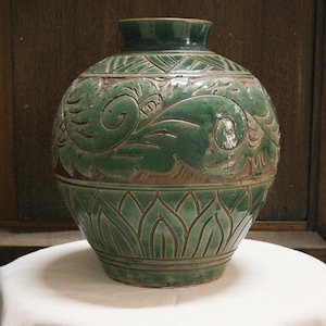 May include: A green ceramic vase with intricate carved floral and geometric patterns. The vase has a rounded body and a narrow neck. The design includes stylized leaves and lotus shapes, with a dark green glaze and a brown underglaze.