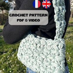 Può includere: Una borsa a uncinetto blu chiaro con un motivo a forma di stella. La borsa ha una tracolla lunga e viene indossata sulla spalla. L'immagine include il testo "CROCHET PATTERN PDF & VIDEO" e bandiere per la Francia e il Regno Unito.
