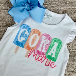 May include: White sleeveless shirt with the name "Gora Marie" printed in colorful popsicle designs. A large light blue bow is placed on top of the shirt. The shirt has ruffle sleeves.