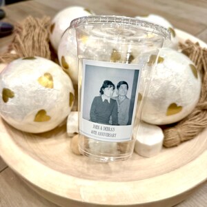 May include: A clear plastic cup with a lid, featuring a vintage photo label that reads "JOHN & DEBRA'S 40TH ANNIVERSARY." The cup sits in a wooden bowl with decorative white spheres adorned with gold hearts and twine.