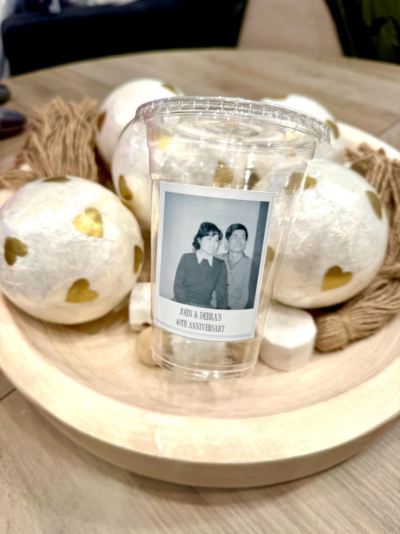 May include: A clear plastic cup with a lid, featuring a vintage photo label that reads "JOHN & DEBRA'S 40TH ANNIVERSARY." The cup sits in a wooden bowl with decorative white spheres adorned with gold hearts and twine.