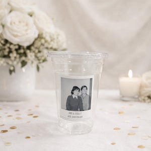 May include: Clear plastic cup with a lid, featuring a black and white photo of a couple. The photo is labeled "John & Debra's 40th Anniversary." The background includes white roses and a lit candle.
