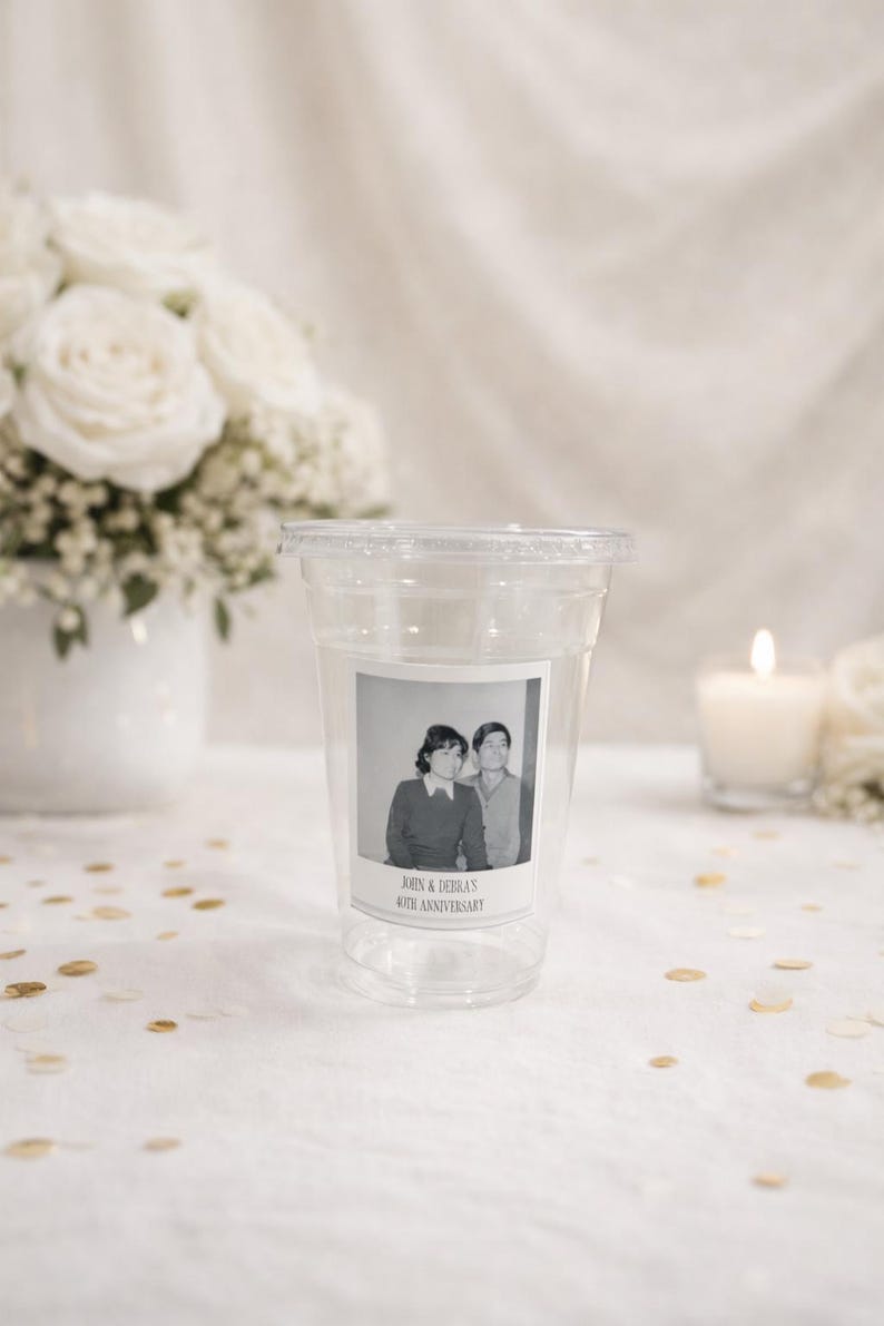 May include: Clear plastic cup with a lid, featuring a black and white photo of a couple. The photo is labeled "John & Debra's 40th Anniversary." The background includes white roses and a lit candle.