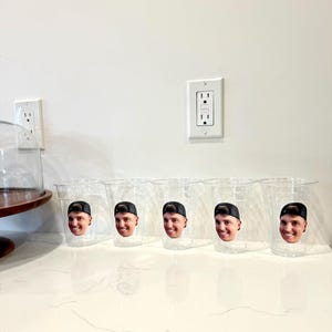 May include: Five clear plastic cups with a smiling face printed on each. The face is wearing a black baseball cap. The cups are arranged on a white countertop.