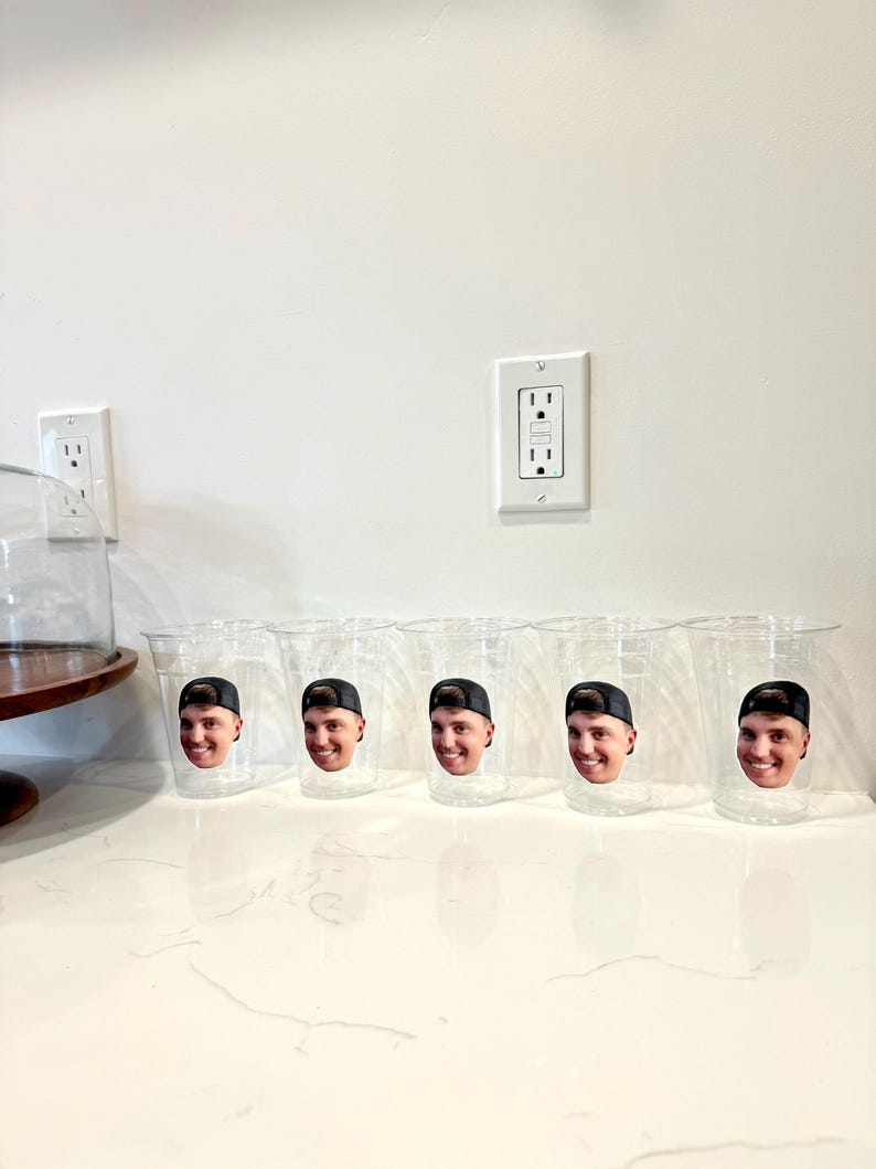 May include: Five clear plastic cups with a smiling face printed on each. The face is wearing a black baseball cap. The cups are arranged on a white countertop.