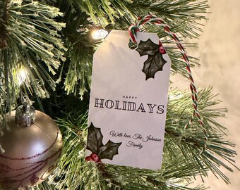 May include: A white gift tag with the words "HAPPY HOLIDAYS" and "With love, The Johnson Family" printed on it. The tag is decorated with holly leaves and red berries. It hangs on a Christmas tree with a gold ornament.