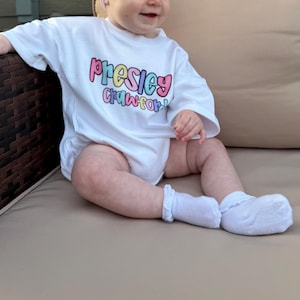 May include: A white baby onesie with the name "Presley Crawford" in colorful bubble letters. The baby is wearing white socks and is sitting on a beige surface.