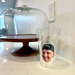 May include: A clear plastic cup with a smiling face sticker on it. A wooden cake stand with a glass dome is in the background. The cup is sitting on a white countertop.