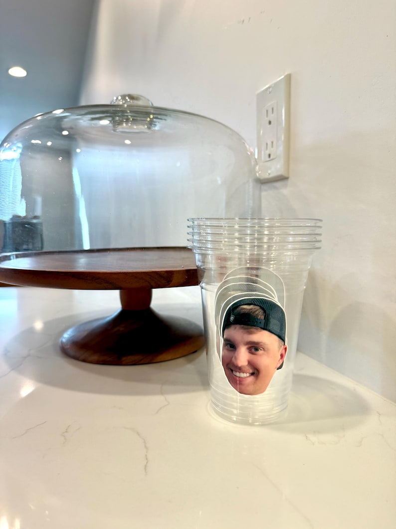 May include: A clear plastic cup with a smiling face sticker on it. A wooden cake stand with a glass dome is in the background. The cup is sitting on a white countertop.