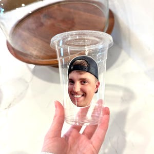 May include: A clear plastic cup with a lid, featuring a sticker of a smiling person wearing a black baseball cap. The cup is held in a hand, with a wooden and glass serving dish in the background.