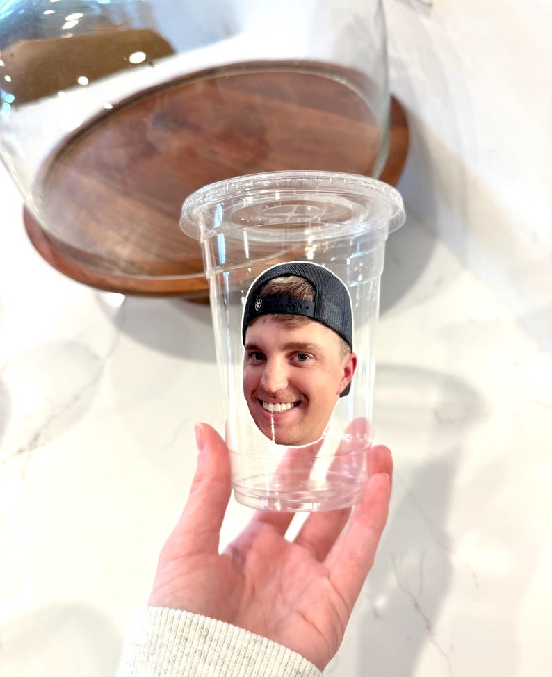 May include: A clear plastic cup with a lid, featuring a sticker of a smiling person wearing a black baseball cap. The cup is held in a hand, with a wooden and glass serving dish in the background.