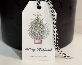 May include: A white gift tag with a watercolor illustration of a Christmas tree, adorned with lights and a pink base. The tag reads "merry christmas" and "LOVE, THE JOHNSONS" in a handwritten font, with a black and white twine.