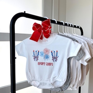 May include: White baby bodysuit with a red, white, and blue fireworks design and the name "Avery Claire" in red script. A large red bow with white trim is attached to the hanger. Other bodysuits in gray and white are also visible.