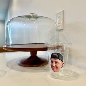 May include: A clear plastic cup with a sticker of a smiling person wearing a black baseball cap. A wooden cake stand with a glass dome is behind the cup. The stand has a dark brown base and a light brown top.