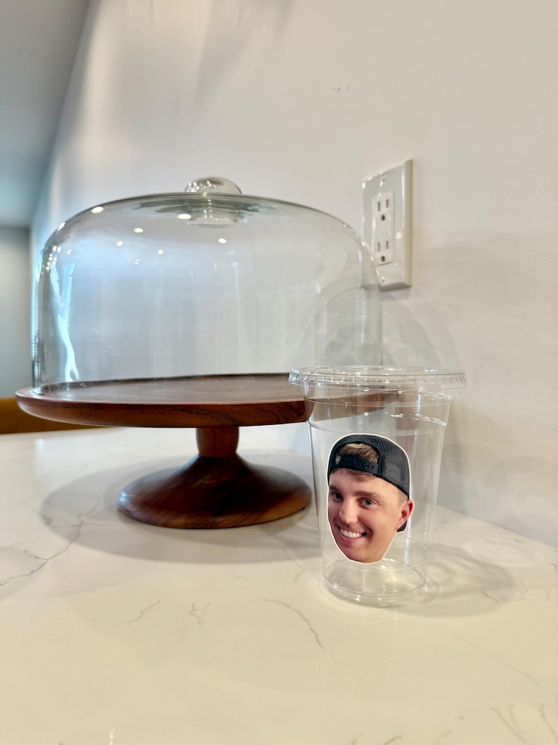 May include: A clear plastic cup with a sticker of a smiling person wearing a black baseball cap. A wooden cake stand with a glass dome is behind the cup. The stand has a dark brown base and a light brown top.