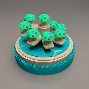 Può includere: Una scatola decorativa rotonda color turchese con una serie di figurine di funghi verdi con pois viola. La scatola ha un bordo beige chiaro e una base turchese con un design decorativo. I funghi sono disposti attorno al centro.