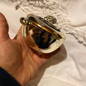 Könnte beinhalten: Eine silberfarbene, herzförmige Schmuckschatulle mit einem kleinen, dekorativen Knopf mit Blumenmuster auf der Oberseite. Die Schatulle hat einen Klappdeckel und einen kleinen, dekorativen Griff mit Blumenmuster. Die Schatulle wird in einer Hand gehalten.