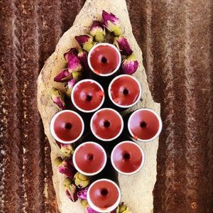 May include: A collection of ten small, dark red, round candles arranged in a pyramid shape on a light brown stone surface. The candles are surrounded by dried pink rose buds.