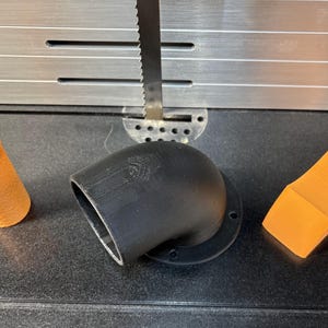 May include: Three 3D-printed objects: an orange handle, a black angled tube, and an orange rectangular piece. The objects are on a dark surface with a metal band saw blade in the background.