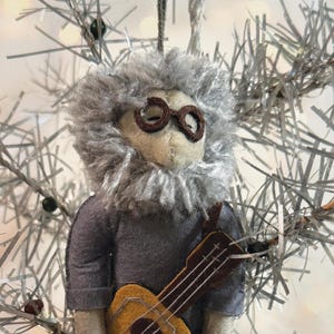 May include: A handmade felt ornament of a musician with a gray beard and glasses, wearing a gray shirt and holding a ukulele. The ornament is set against a backdrop of silver tinsel.