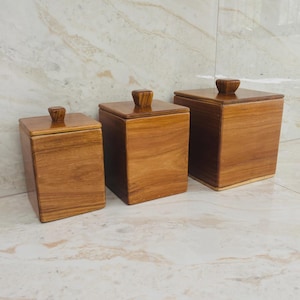 May include: Three wooden storage containers of varying sizes. Each container is square with a flat lid and a small, rounded knob. The wood grain is visible, and the color is a warm, medium brown. The containers are arranged in a row.