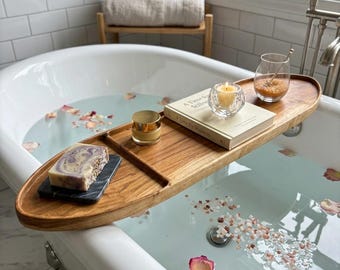 Puede incluir: Una bandeja de madera ovalada para bañera sobre una bañera blanca con patas, llena de agua, pétalos de rosa y sales de baño. La bandeja sostiene una pastilla de jabón veteado sobre un posavasos de piedra negra, un tarro dorado, un libro y una vela.