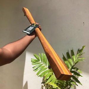 May include: A long wooden paddle with a rectangular handle and a wide, flat blade. The paddle is made of light and dark brown wood, with a smooth finish. A tag with a QR code is attached to the handle. The paddle is held in front of a white wall and green plant.