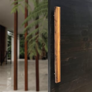 May include: A vertical wooden door pull with a natural wood grain finish. The door pull is rectangular and attached to a dark, possibly black, surface. The wood tones range from light to dark brown.