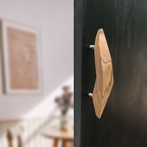 May include: A wooden door handle with a geometric design. The handle is made of light brown wood and is attached to a black door. The handle has a unique, angular shape and is mounted with silver hardware.