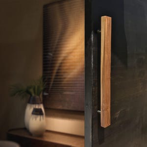 Könnte beinhalten: Ein rechteckiger Türgriff aus Holz mit natürlicher Holzmaserung. Der Türgriff ist an einer dunkel gefärbten Tür montiert. Der Griff ist etwa 30 cm lang und mit silberner Hardware befestigt. Die Tür ist teilweise geöffnet und gibt einen Einblick in den Innenraum.