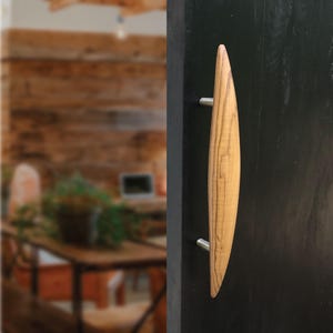 Könnte beinhalten: Ein Türgriff aus Holz mit hellbraunem Finish und geschwungenem, länglichem Design. Der Türgriff ist mit zwei silberfarbenen Schrauben an einer schwarzen Tür befestigt. Der Hintergrund ist unscharf und zeigt einen Blick in einen Raum.