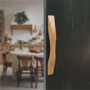Könnte beinhalten: Ein helles Holzmöbelgriff mit geschwungener, ergonomischer Form. Der Griff ist an einer dunklen, möglicherweise schwarzen Schranktür befestigt. Der unscharfe Hintergrund zeigt eine Küche mit Tisch und Stühlen.