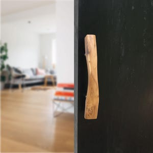 Könnte beinhalten: Ein Türgriff aus hellem Holz mit geschwungener Form, montiert an einer schwarzen Tür. Der Griff zeigt die natürliche Maserung des Holzes. Befestigt mit silbernen Schrauben. Der Hintergrund ist unscharf.