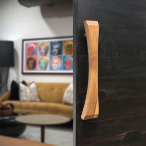 Puede incluir: Un tirador de puerta de madera con un diseño curvo. El tirador es de madera clara y está montado en una puerta de madera teñida oscura. El fondo muestra un salón con una obra de arte colorida y un sofá color mostaza.
