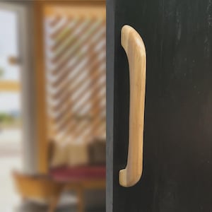 May include: A light-coloured wooden door handle with a curved design is mounted on a dark surface. The handle has a smooth, polished finish and a simple, modern aesthetic. The background is blurred, suggesting an interior setting.