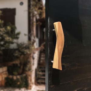 Könnte beinhalten: Ein hölzerner Türgriff mit einem einzigartigen, geschwungenen Design. Der Griff besteht aus zwei Holztönen, hell und dunkel, und ist mit silbernen Beschlägen an einer dunklen Tür befestigt. Die Form des Griffs ist organisch und modern.